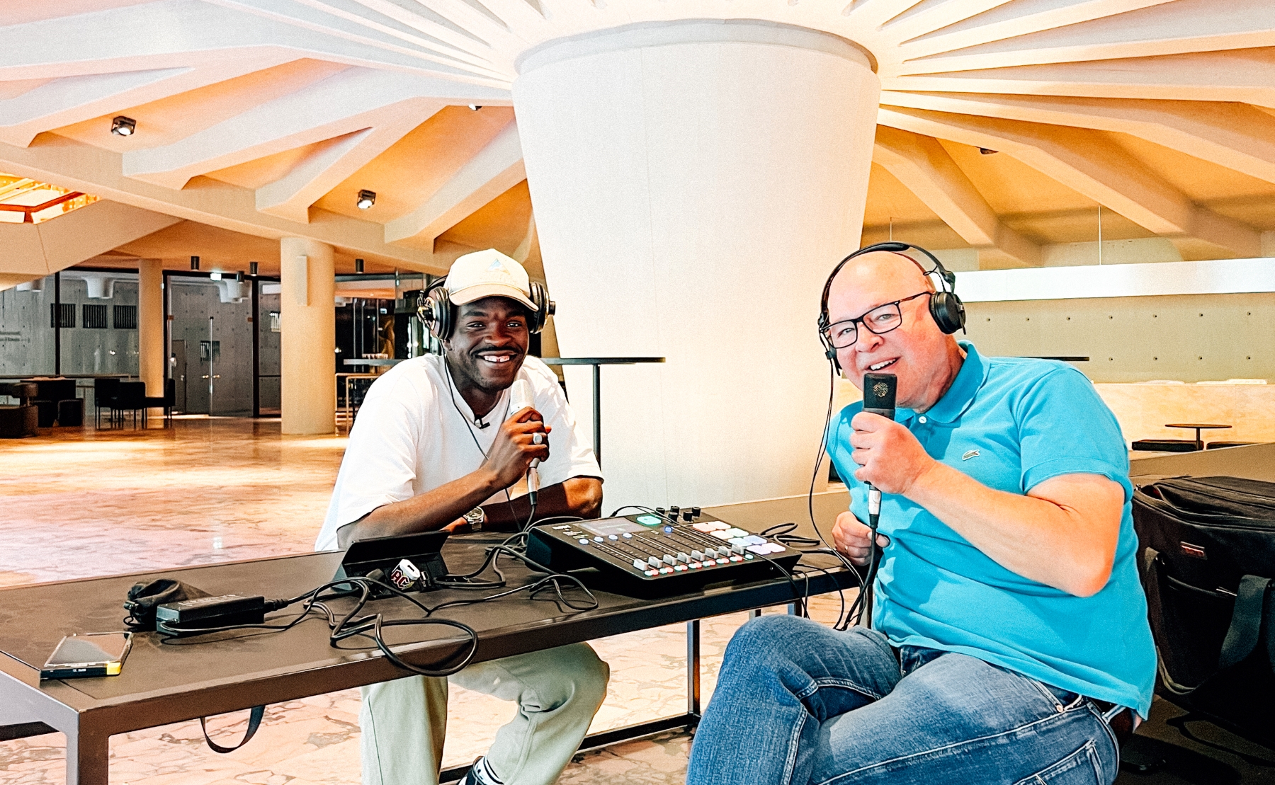 Flockey Ocscor und Mike Litt sitzen im Foyer des Düsseldorfer Schauspielhauses an einem Tisch mit Aufnahmegeräten und blicken lachend in die Kamera.