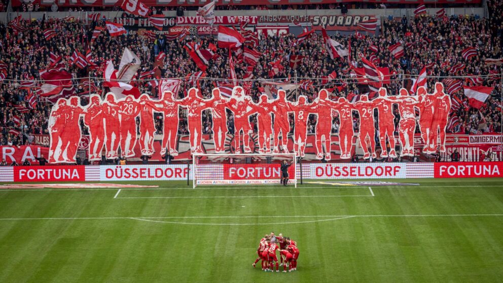 "Glaub dran -Gebt alles!" Steht auf einem riesengroßen Banner über der letzten Reihe im Düsseldorfer Fußballstadion. Man sieht außerdem rotweiß gekleidete Fans und auf dem Rasen die Mannschaft im Kreis stehend.