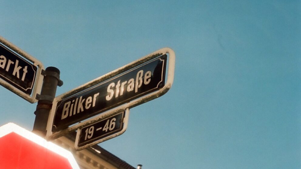 You can see a cropped stop sign, in the background pink old buildings, the street sign Bilker Straße and blue sky