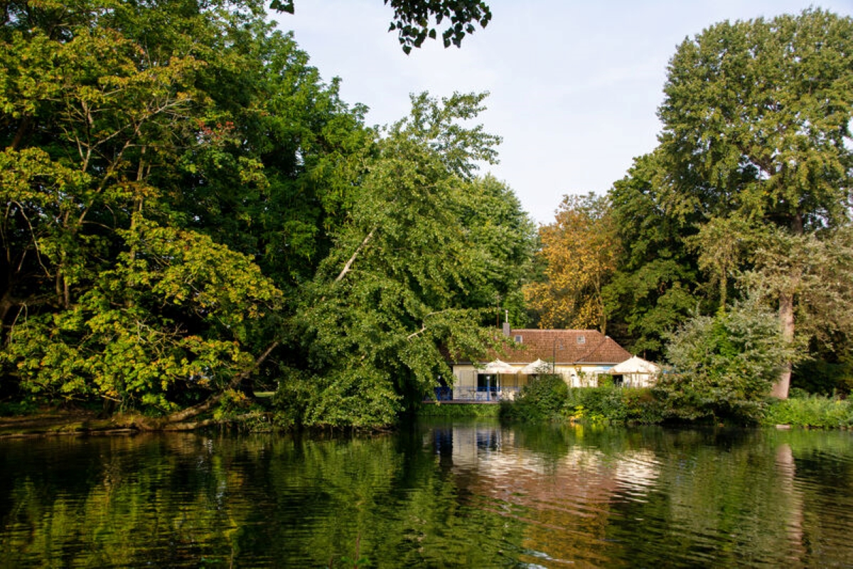 The boathouse in the south park can be seen in the background, in front of it a lake framed by tall trees.