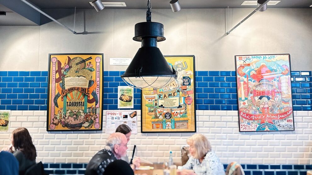In the Takumi restaurant, very colorful poster prints hang in frames on a blue and white tiled wall, in front of which you can see guests eating at a table.