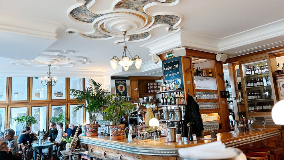 Französische Küche serviert auch das Robert. am Rheinufer. Man sieht den Innenraum mit einer typisch französischen Bar und Theke aus dunlem Holz, einen Jahrhundertwendeleuchten der Mitte der Stuckverzierten Decken und im Hintergrund große Fenster und Bistrotische.
