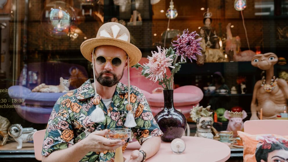 Rolf Buck trägt einen Overall mit bunten floralen Prints, einen melonenartigen Strohhut eine runde Hornsonnenbrille und hält ein Cocktailglas in der Hand, während er vor dem Schaufenster seines Stores Vaseline sitzt.