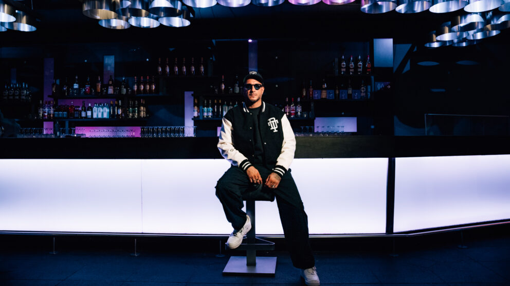 Loco Dice, DJ, sitzt auf einem Barhocker, hinter ihm die weiß angeleuchtete Bar.