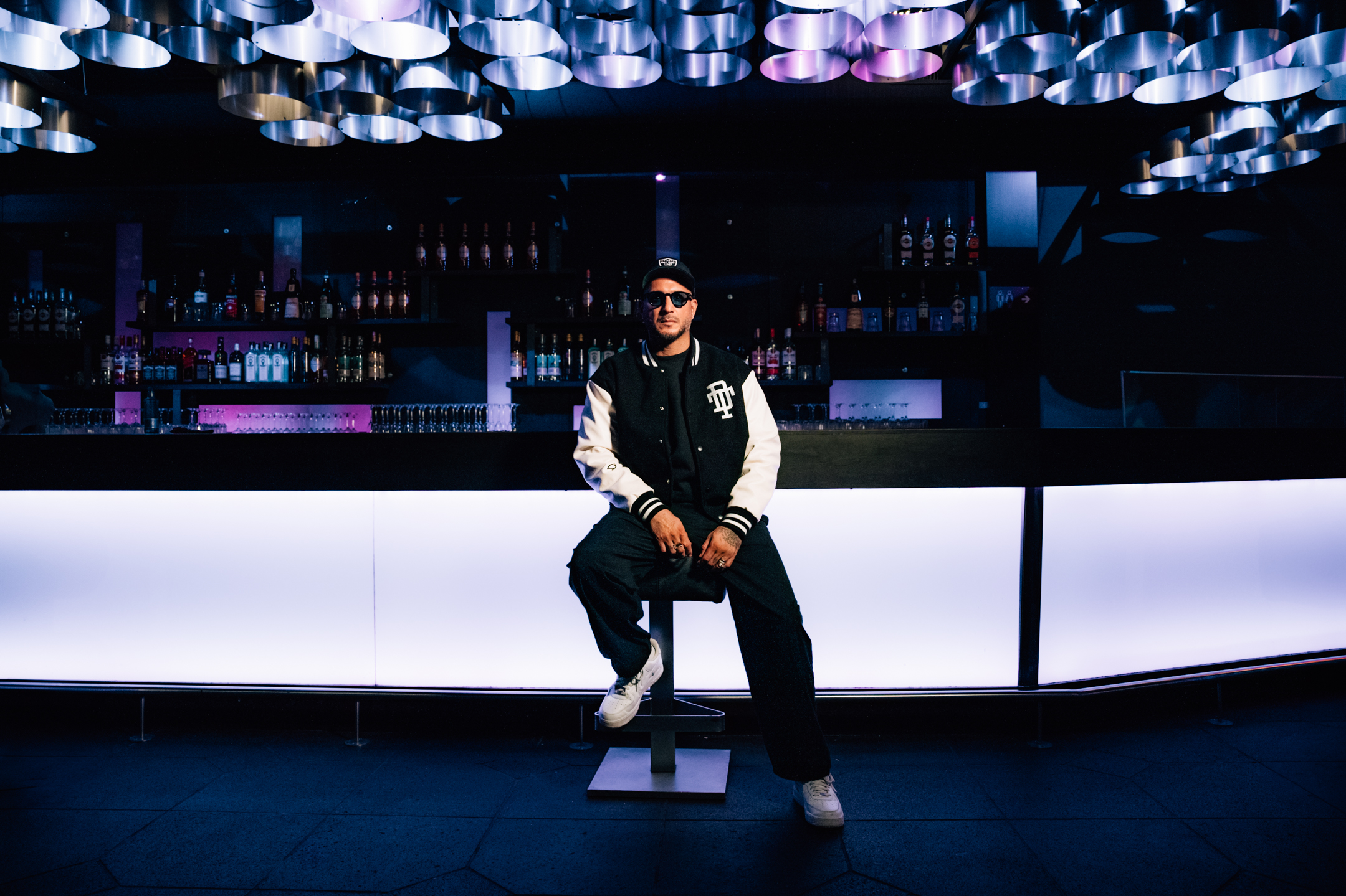 Loco Dice, DJ, sitzt auf einem Barhocker, hinter ihm die weiß angeleuchtete Bar.