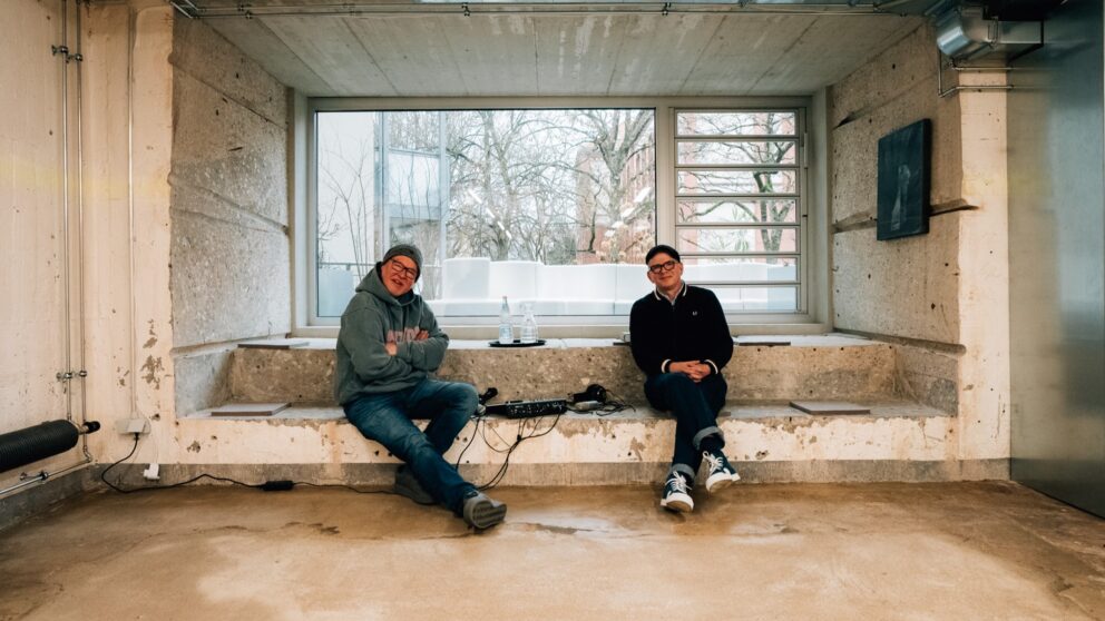Mike Litt (links) und Tobi Dahmen sitzen im Ausstellungsraum im Bilker Bunker, hinter ihnen ein großes Fenster.