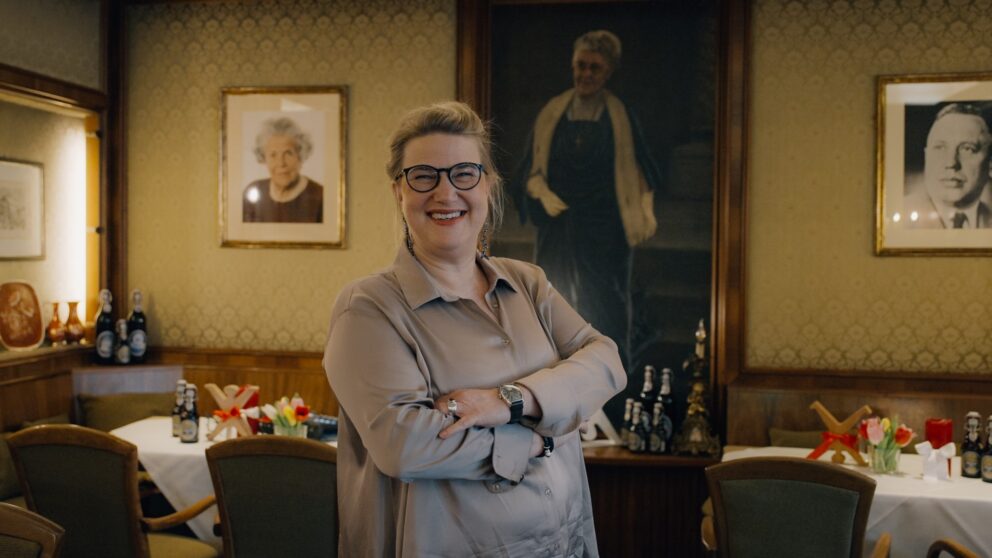 Thea Ungermann, Chefin der Brauerei Schumacher, steht in einem Raum der Hausbrauerei. Im Hintergrund sieht man Portraits und eingedeckte Tische.