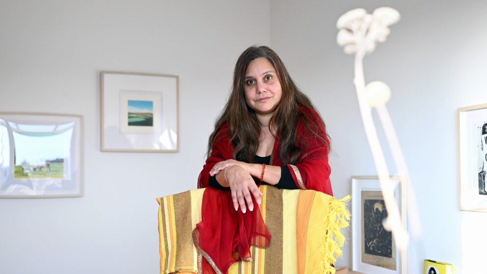 Writer Mithu Sanyal stands leaning slightly forward, her hands resting on the back of a chair with a yellow blanket on it.
