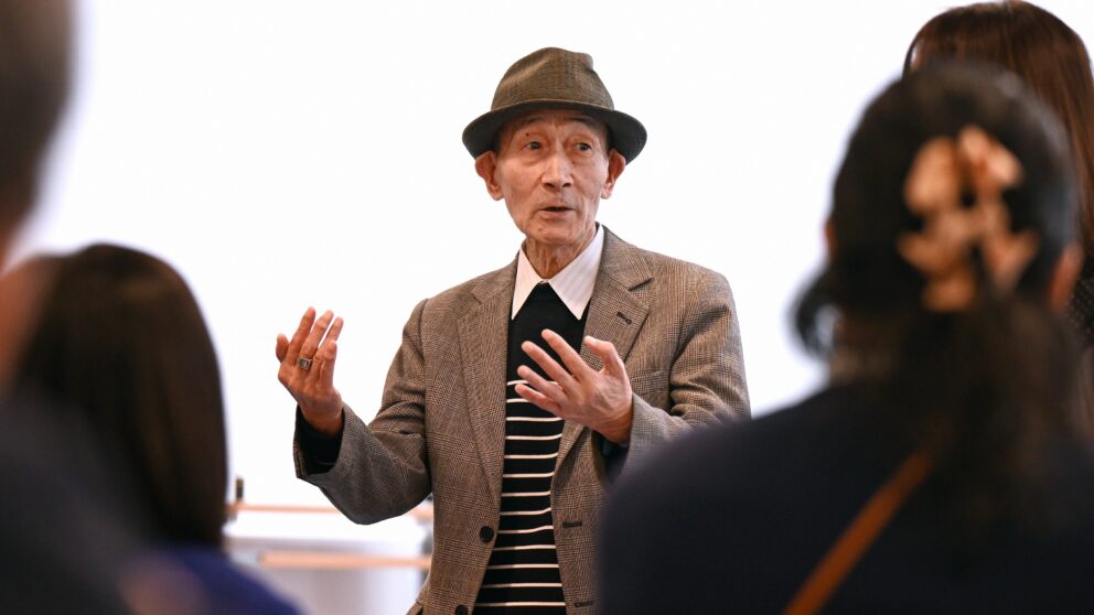 You see an elderly Japanese man with a hat standing in front of a choir.