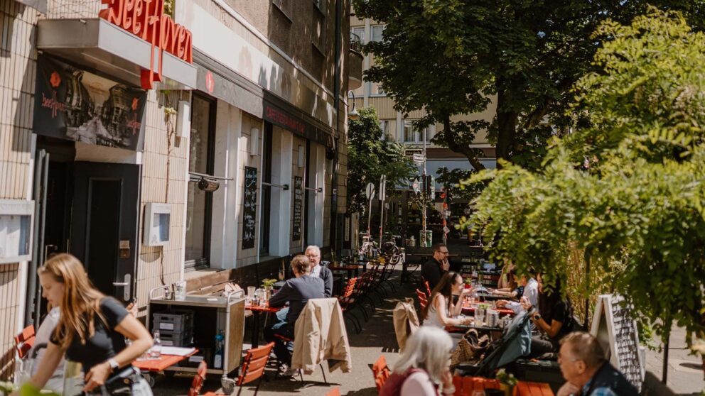 Man sieht einen Biergarten, in dem Menschen sitzen.