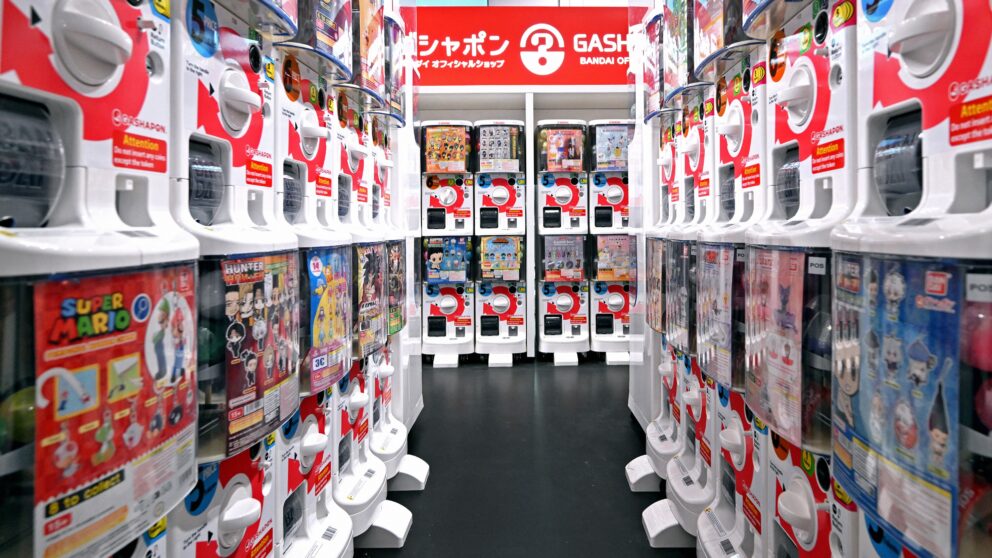 Man sieht einen Gang aus Gashapon-Automaten. Das sind Automaten, die ähnlich wie früher Kaugummiautomaten aussehen. Oben sind sie transparent und man sieht Kapseln mit Spielzeug, darunter kann man eine Plastikmünze einwerfen und durch Drehen eines Drehschalters fällt dann eine Kapsel raus.